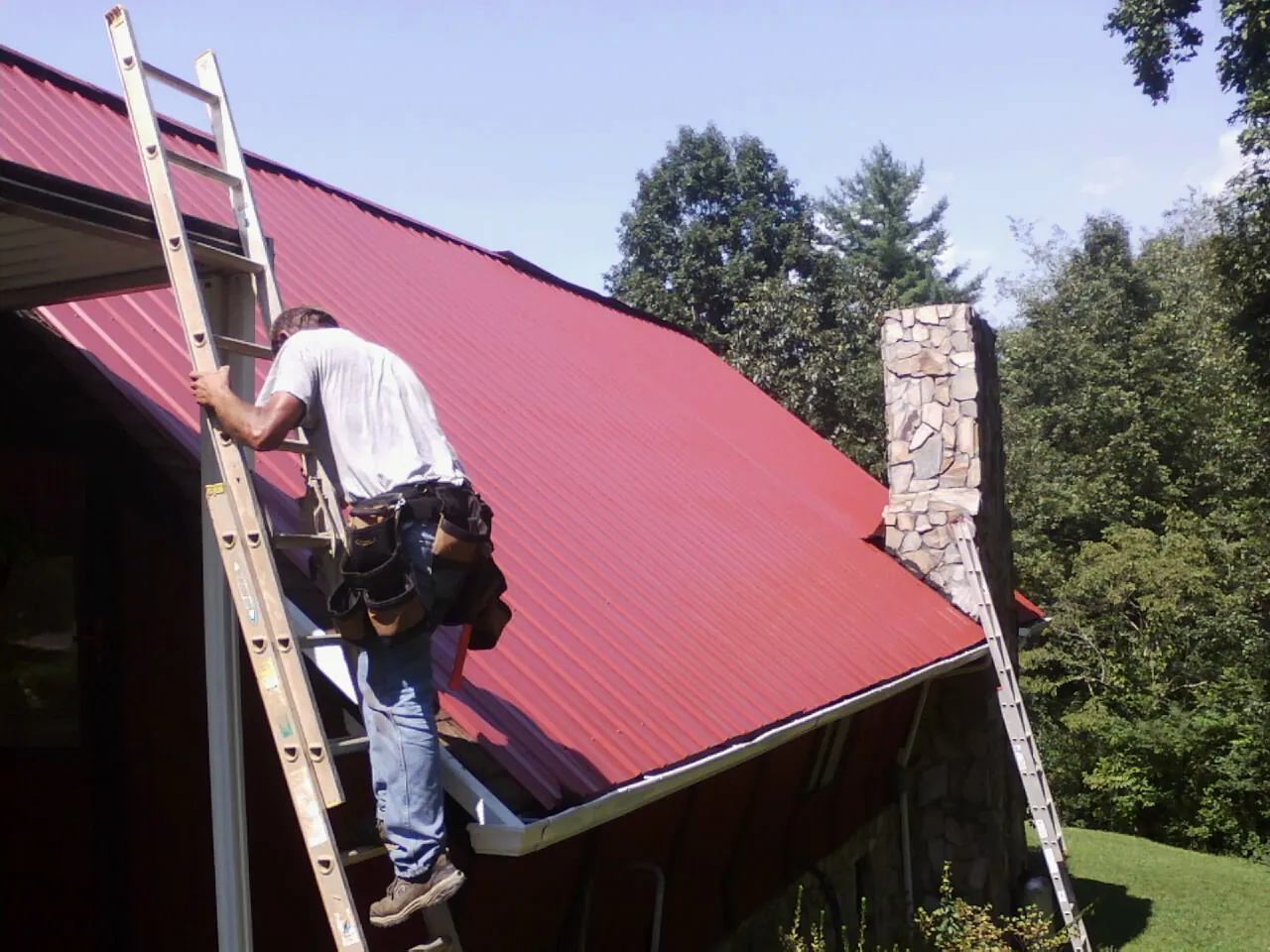 Roofer on a ladder beside a red metal roof and stone chimney — Soffit & Fascia Repair by Roofing Brothers in Greenback